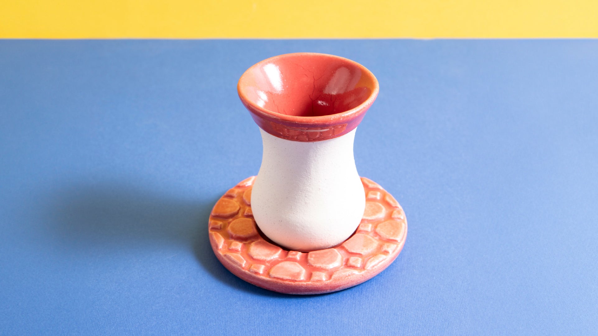 Singular view of a red and white cup with its ceramic red coaster, blue and yellow background